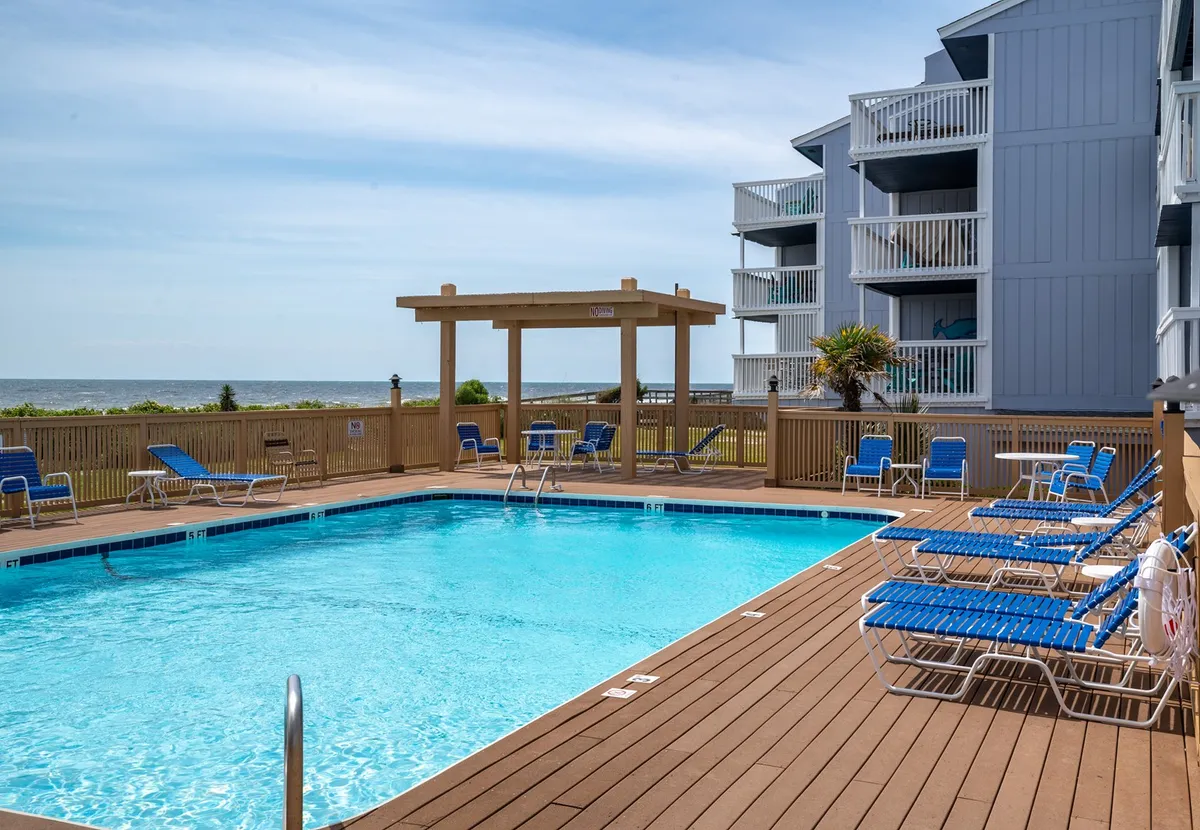 This exterior image features a swimming pool with lounge chairs, a shaded pergola, and an ocean view in the distance. The inviting setting highlights opportunities for sunbathing, swimming, and tranquil poolside moments, especially in-season.