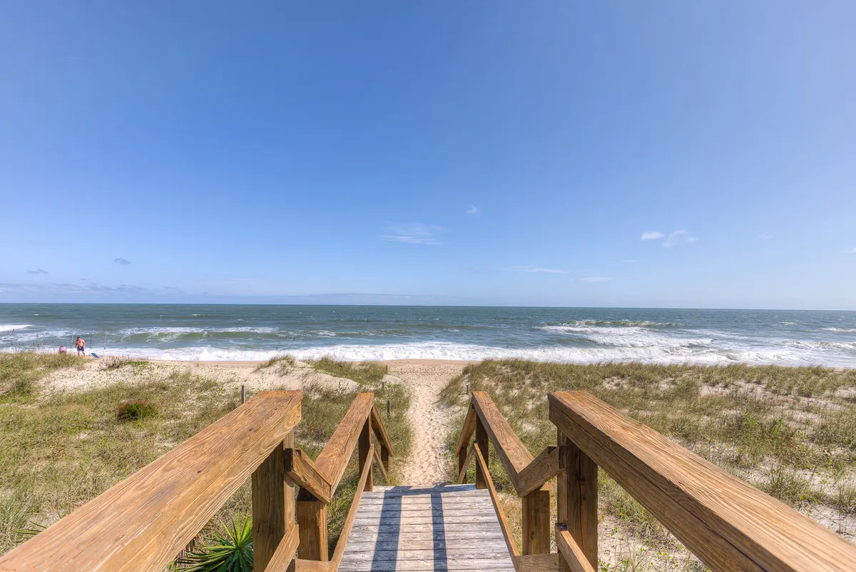 A wide wooden walkway stretches over soft dunes and leads directly to the sandy beach, bordered by dune grasses and framed by gentle ocean waves in the distance. The path evokes a sense of anticipation and the promise of easy beach access.