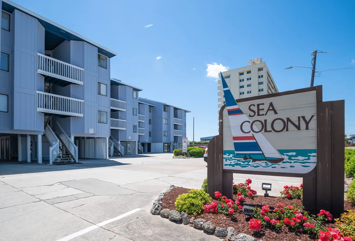 A welcoming view of the 'SEA COLONY' sign with modern apartment buildings in the background and beautifully landscaped flowers in the foreground, suggesting a friendly, vibrant community atmosphere.