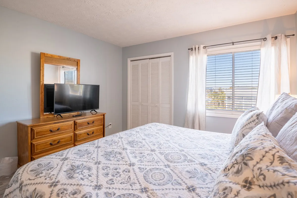 Another angle of the bedroom showcases a bed adorned with decorative pillows, a dresser with a mirror for ample storage, a television, and a sunlit window. The ambiance is serene and restful, offering both comfort and functionality for longer or shorter stays.