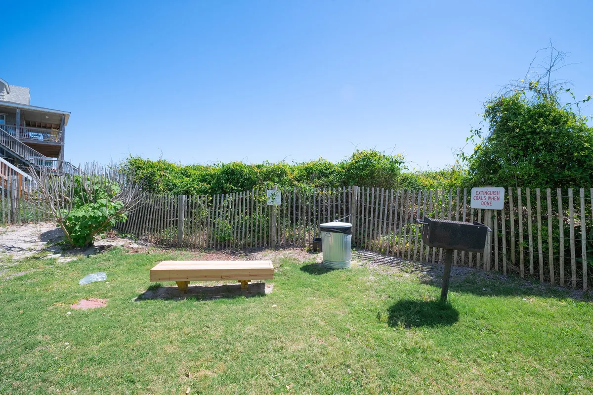 A casual picnic area featuring a sturdy bench and a grill, set amidst light vegetation and bordered by a fence. The space is ideal for laid-back barbecues, picnics, and enjoying fresh air with loved ones.