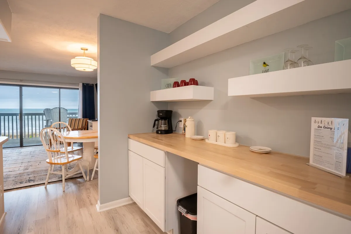 A cozy dining area anchored by a wooden dining table, with comfortable chairs, a coffee maker on the counter, and shelves neatly stacked with dishes. The scene is warm and welcoming, ideal for family breakfasts or morning coffee as mentioned in the content.