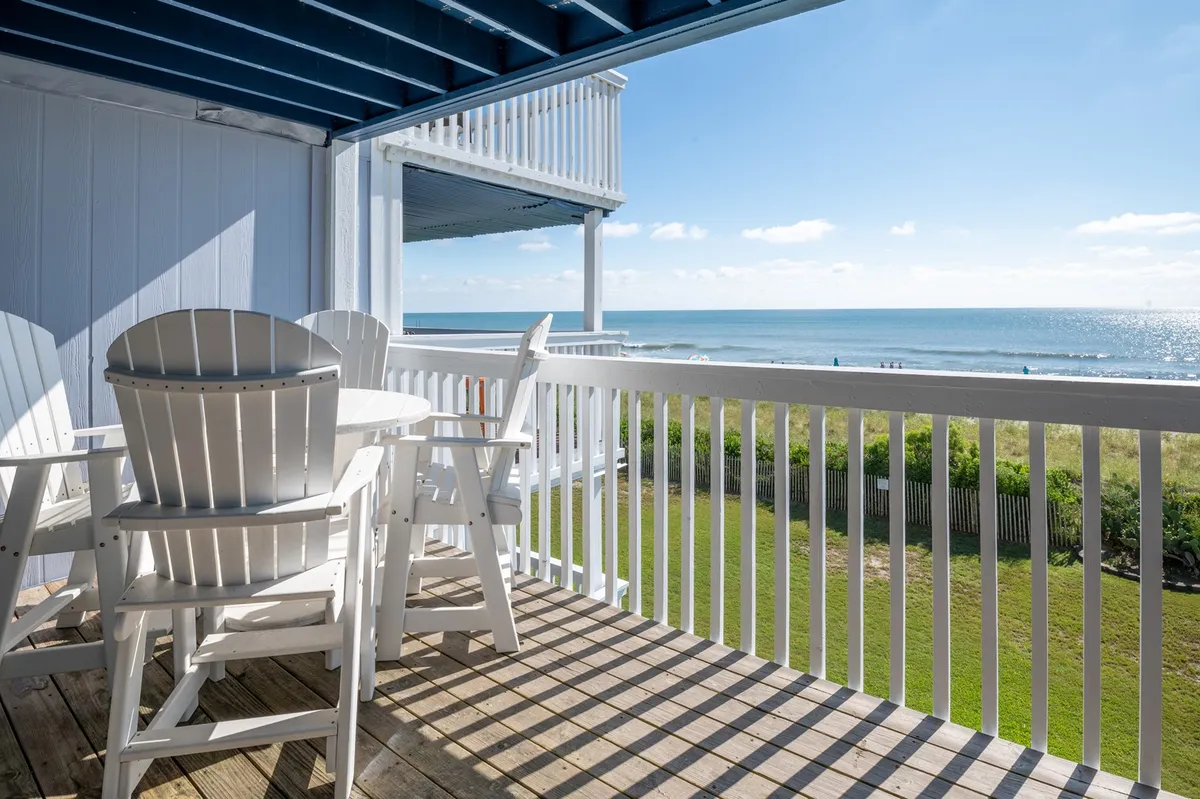 The image shows an outdoor dining table set on a private balcony overlooking the sparkling ocean at Carolina Beach. The view stretches to the horizon, with gentle waves rolling onto the sandy shoreline just beyond the deck. The table is positioned perfectly for enjoying meals or drinks while taking in the tranquil beachscape. The scene exudes relaxation, with the promise of sunrise breakfasts or sunset gatherings.
