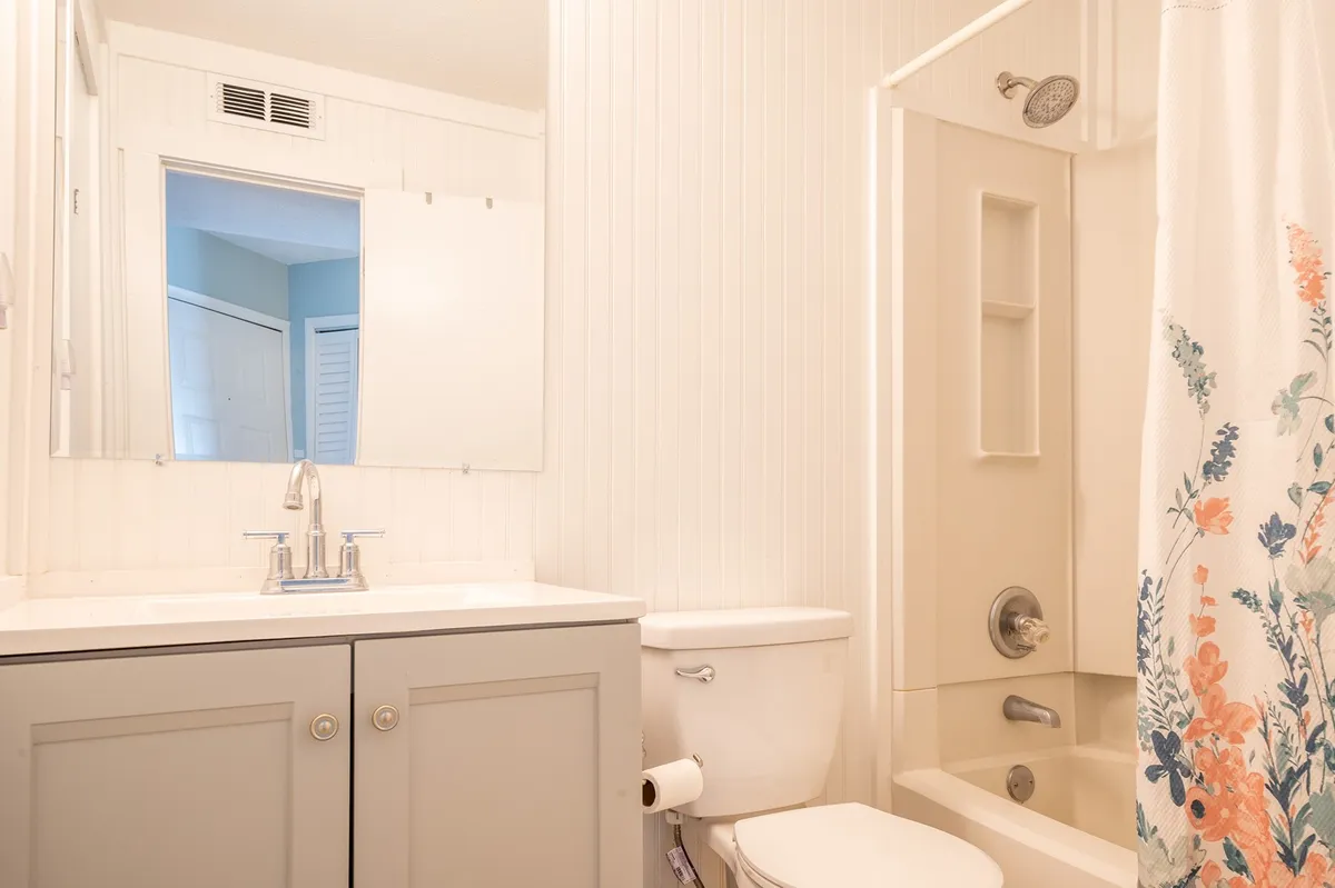 A fresh, sparkling ensuite bathroom featuring a modern sink and faucet, a floral shower curtain, a large mirror, and essential amenities. The bathroom exudes cleanliness, bright lighting, and a sense of refreshing tranquility, perfectly suiting both quick routines and moments of relaxation.