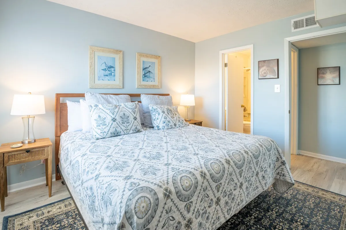 A serene bedroom features a neatly made bed with soft linens, modern artwork above the headboard, a stylish nightstand with a lamp, and gentle natural light streaming through the window. The flooring appears recently updated, and the entire space evokes a calm, inviting retreat perfect for rest or reading.